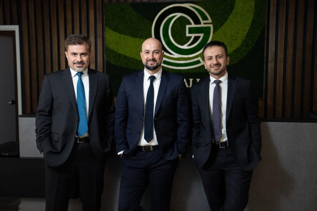 The three founding partners of Onal Gallant law firm, dressed in dark business suits and ties, standing together in front of the firm's logo — a circular 'G' emblem set against a green moss wall in their office