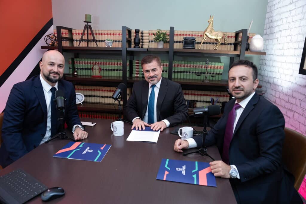 Two Onal Gallant partners seated at a podcast recording table with a guest, surrounded by Shure microphones, Onal Gallant branded mugs and folders, and a bookshelf lined with law books and legal décor including a golden horse statuette and a gavel